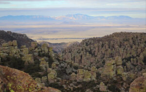 View across a valley with distant mountains on the far side. In the foreground are rock pinnacles and spires. The Chiricahua Mountains are one of many sky islands stretching across southern Arizona. From this view, the Dragoon Mountains on the other side of Sulphur Springs Valley also rise up like an island from the sea of grass.