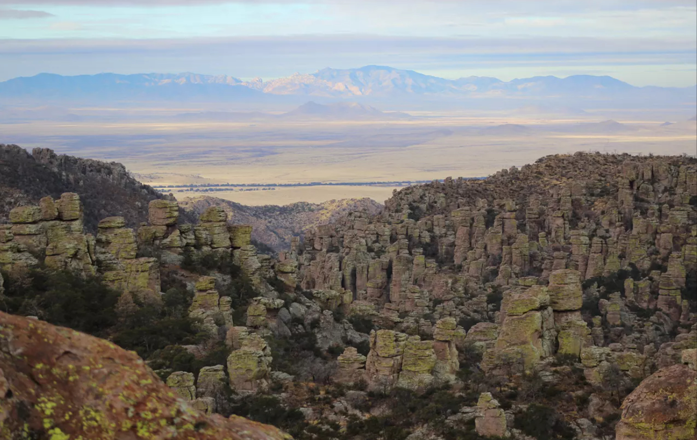 View across a valley with distant mountains on the far side. In the foreground are rock pinnacles and spires. The Chiricahua Mountains are one of many sky islands stretching across southern Arizona. From this view, the Dragoon Mountains on the other side of Sulphur Springs Valley also rise up like an island from the sea of grass.